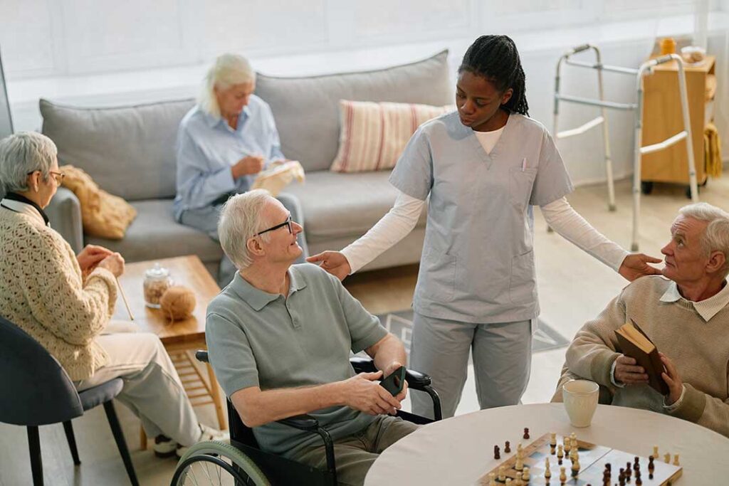 Four older adults smiling and playing cards together at a brightly lit activity table.